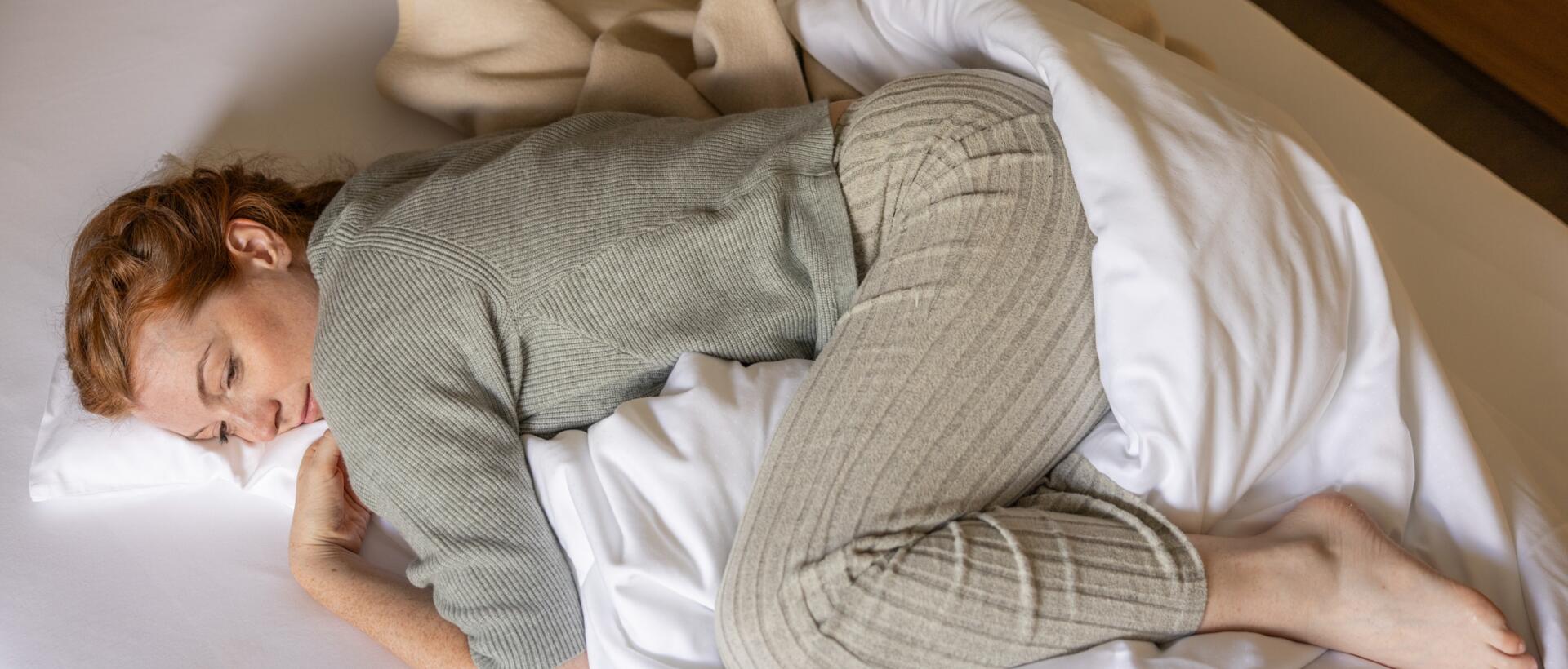 Frau in Loungewear zusammengerollt im Bett – Rückzug und Ruhe bei Regelschmerzen