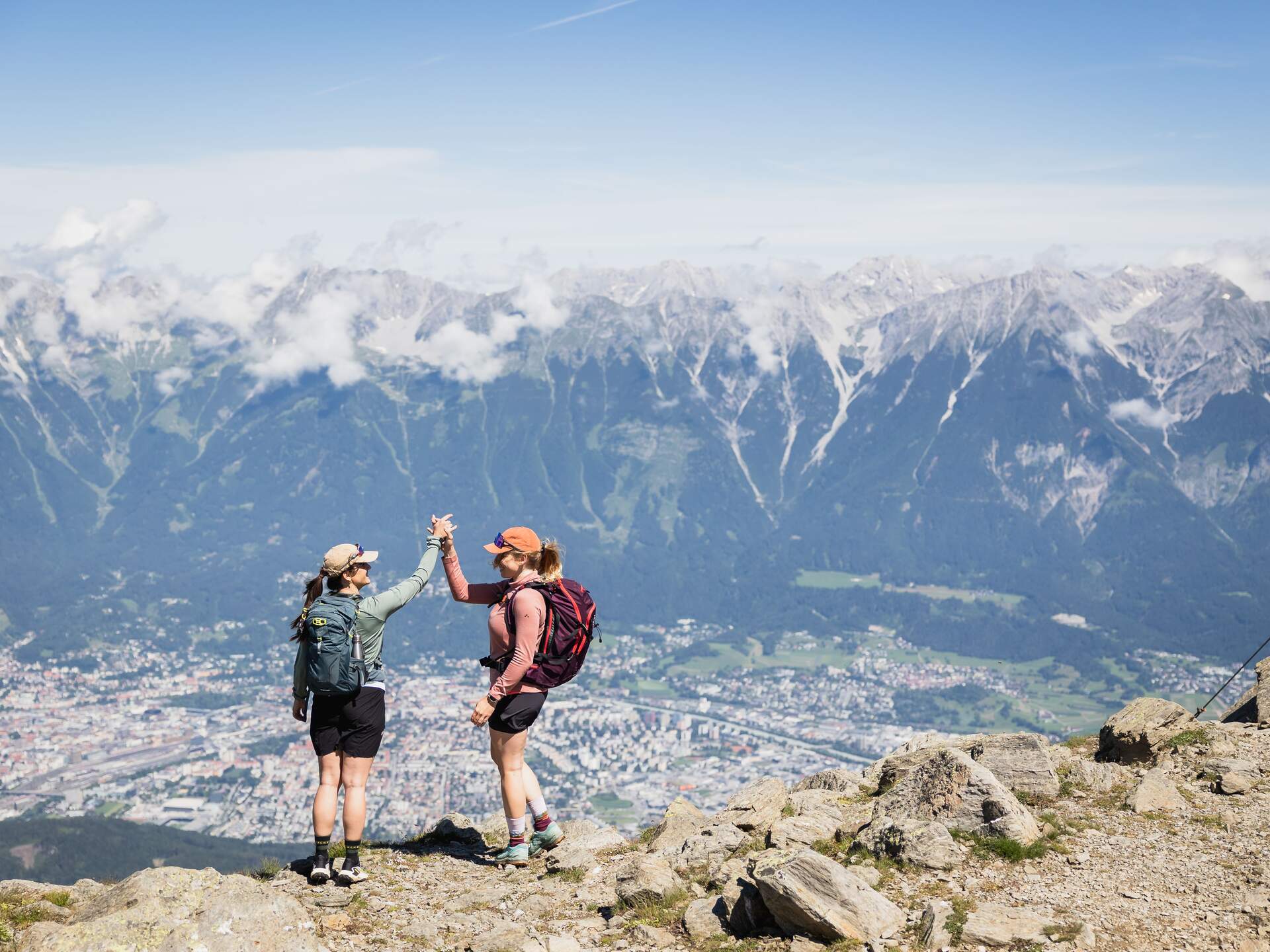 Zwei Wanderinnen geben sich High-Five auf einem Berggipfel mit Blick auf eine Stadt und schneebedeckte Berge | © Jonas Schwarzwälder Photography