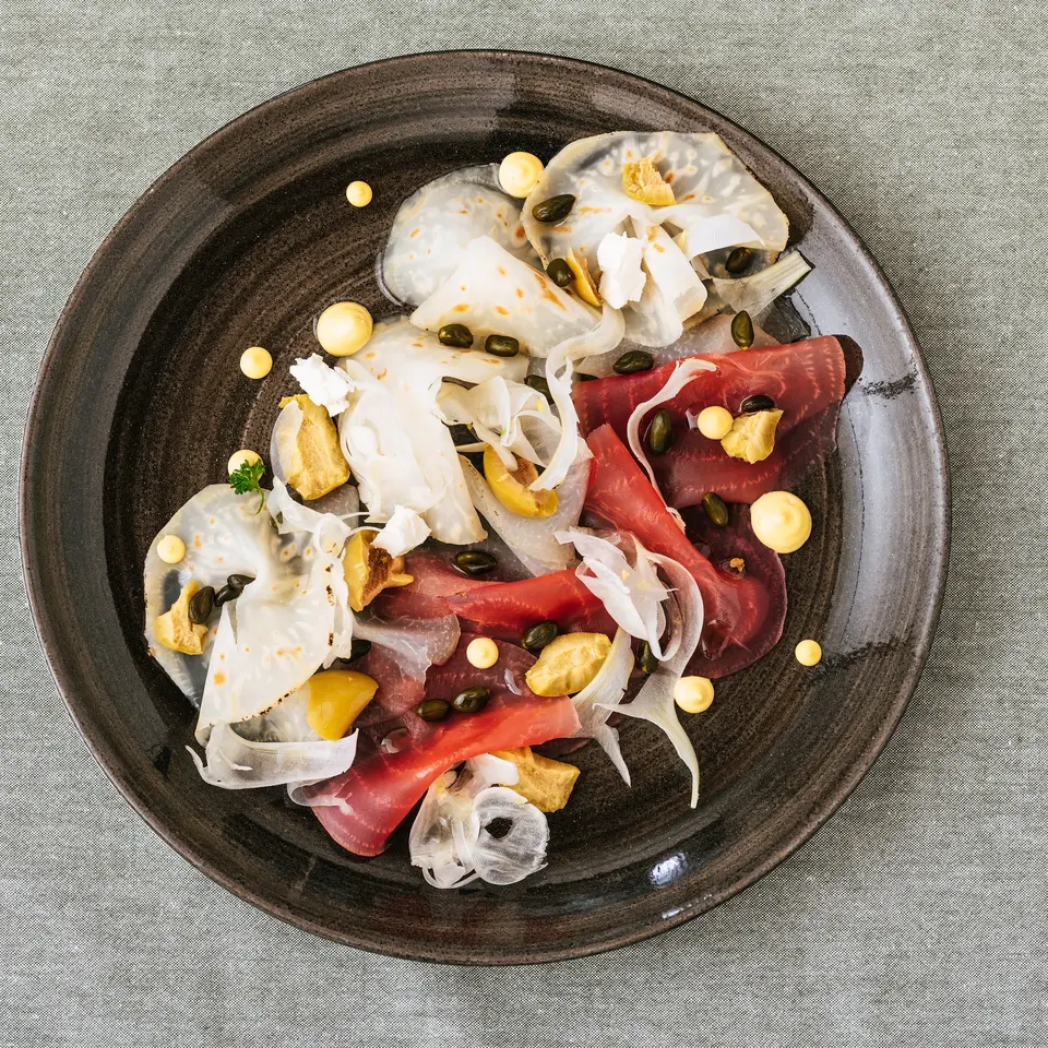 Carpaccio mit Fenchel, Kapern und gelben Tupfen auf dunklem Teller