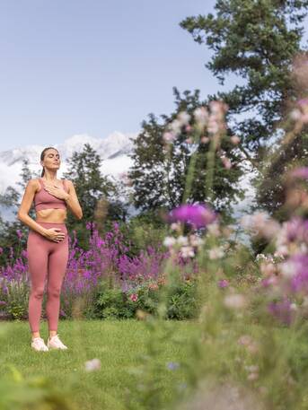 Frau in Sportkleidung meditiert entspannt im Garten mit Blumen und Bäumen vor Bergkulisse